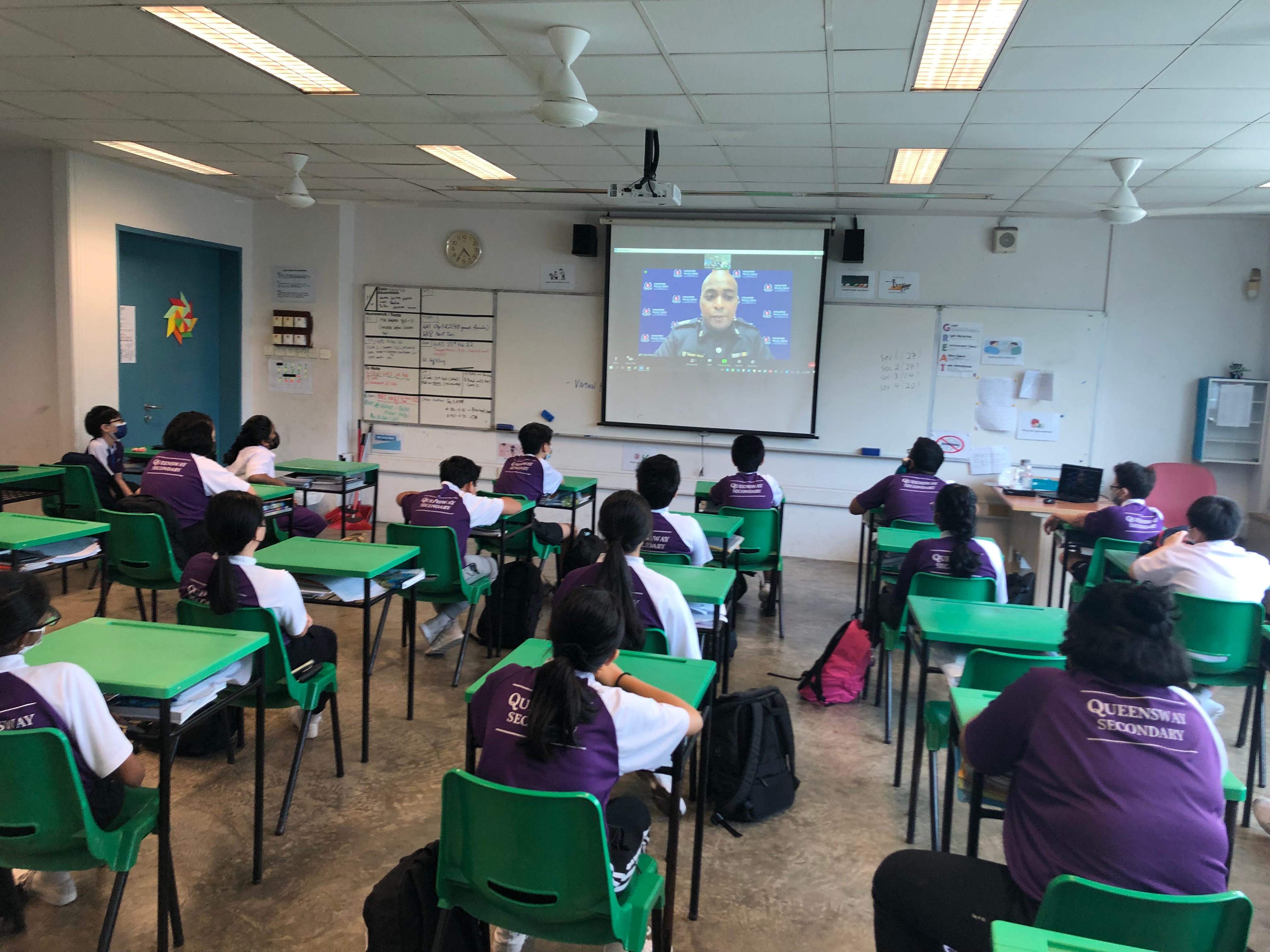 Queensway Secondary students in purple uniforms sit at green desks watching a video call projected on a screen.